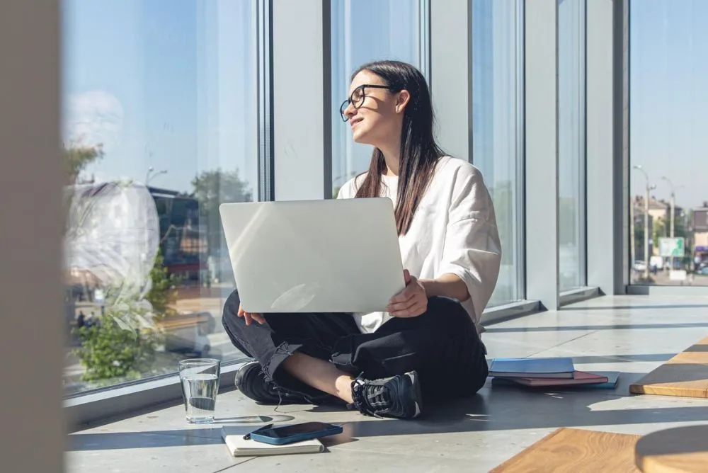 Freelancer sitting cross-legged with a laptop in a bright coworking space for freelancers by a window.