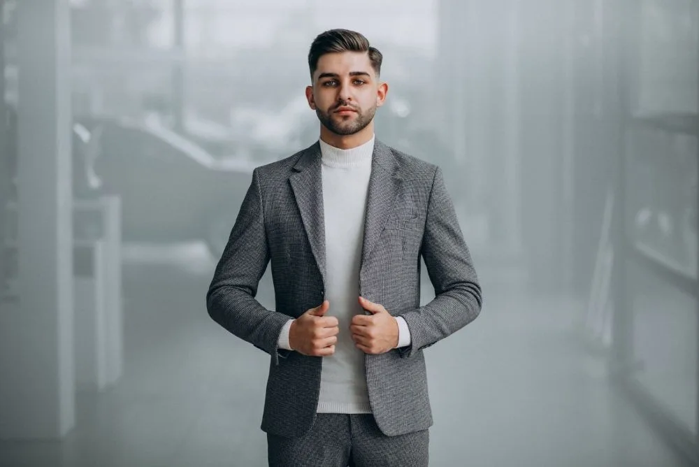 Professional man in a grey suit and white turtleneck standing in a modern, minimalist office setting.