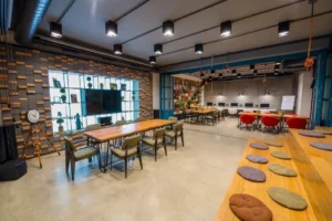 Spacious coworking space with long wooden tables, colorful seating, industrial lighting, and tiered wooden benches.
