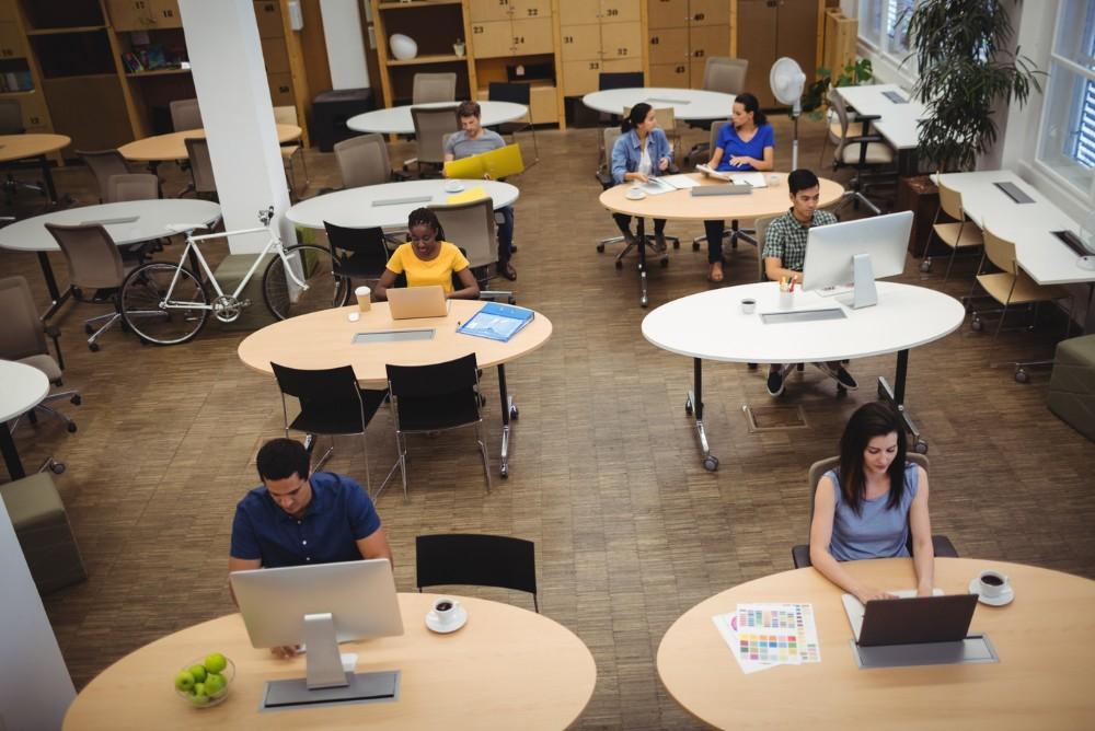 Diverse professionals working at oval tables in a bright, modern coworking space with a bicycle and lockers.