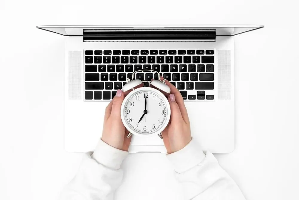 Overhead view of hands holding a white alarm clock over a laptop, symbolizing productivity and timing.