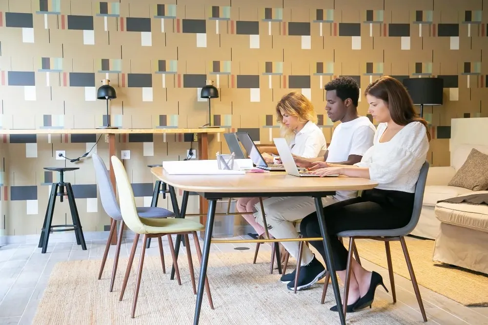 Three remote workers sitting at a wooden table with laptops, collaborating in a stylish, modern common area.