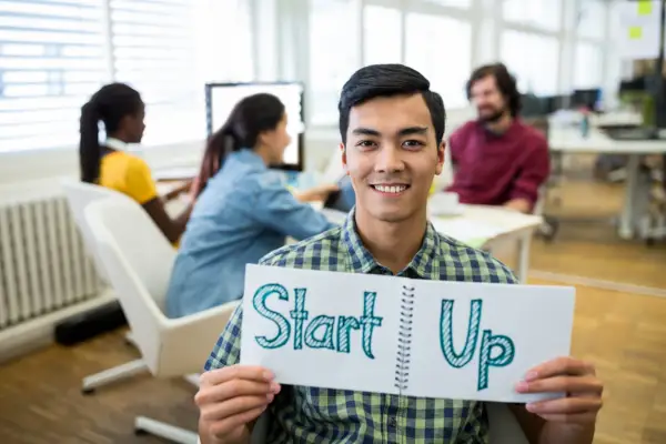 A smiling young man holding a notebook that says "Start Up" in a vibrant office environment, highlighting the benefits of coworking spaces for startups.