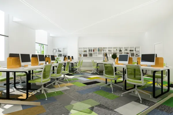 A modern, brightly lit coworking space interior featuring sleek white desks, green ergonomic chairs, computer monitors, and geometric-patterned carpet.
