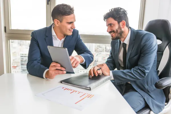 Two businessmen in formal suits sitting at a desk next to a window, smiling and discussing data on a digital tablet during a mentoring session.