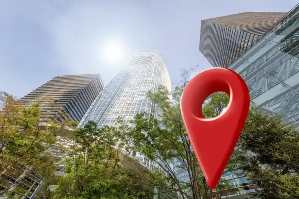 A large red map pin icon positioned in front of modern, glass skyscrapers and green trees against a bright, sunny sky.