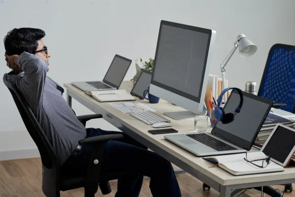 A man relaxes comfortably in a chair at his organized office desk equipped with multiple computer monitors.