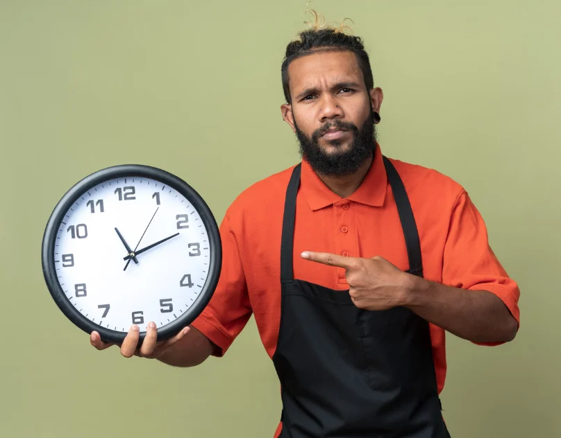 A man in a red polo shirt and black apron looks directly forward while pointing to a large round clock in his hand