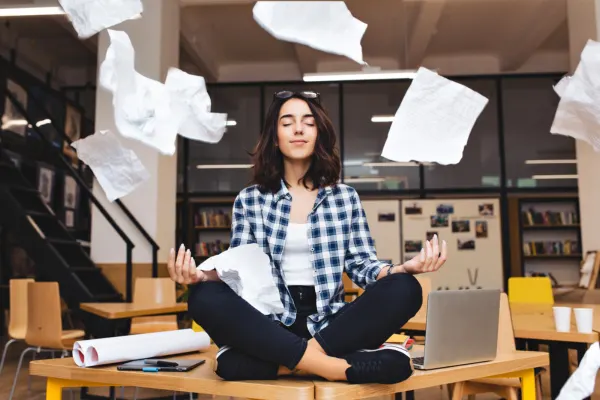 A calm young woman sitting cross-legged on an office desk, meditating peacefully as paper flies all around her.